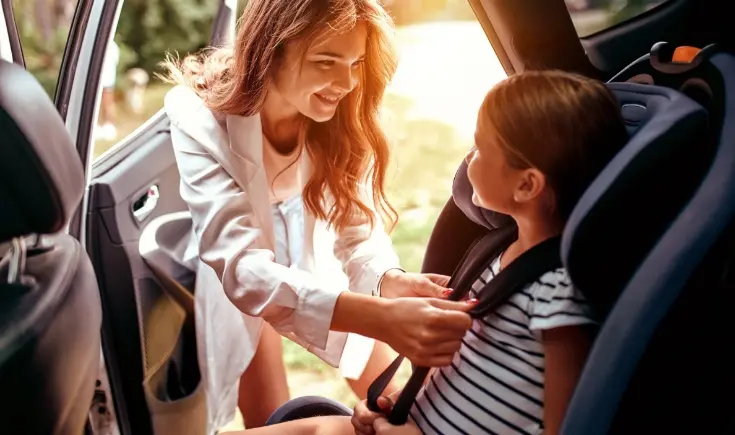 a smiling woman buckling a young girl into a car seat