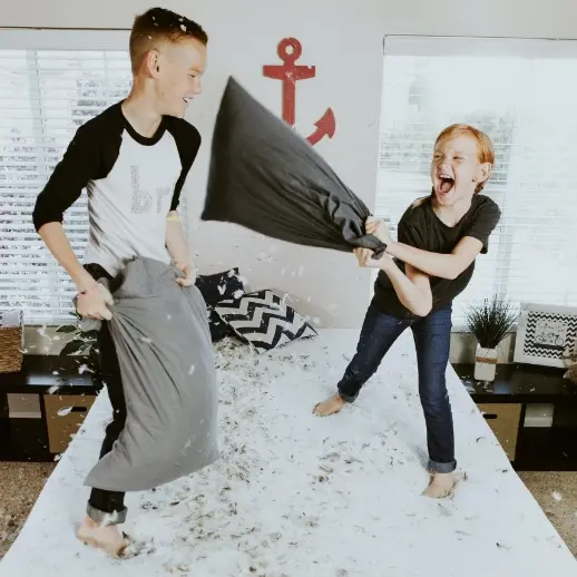 two young kids pillow fighting on a bed, with feathers flying all around them