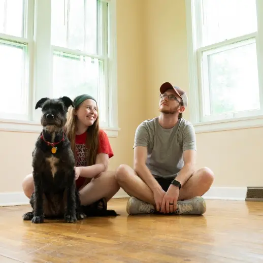 two people and a black dog, sitting on the floor of an empty room