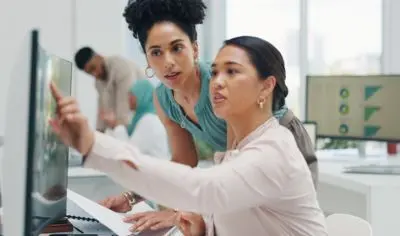 a person pointing to a computer screen, showing someone next to her something