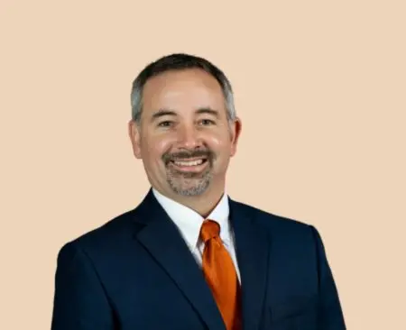 man smiling wearing a navy suit jacket over a white button-up shirt with an orange tie