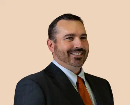 man smiling wearing black suit jacket over a light blue button-up shirt with orange tie