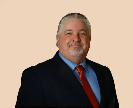 man smiling wearing black suit jacket over a light blue button-up shirt with orange tie