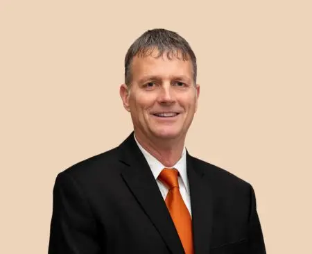 man smiling wearing black suit jacket over a white button-up shirt with orange tie