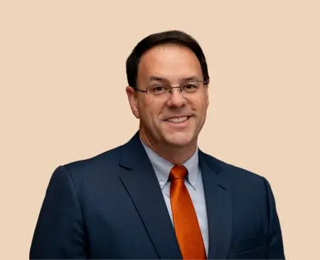 man smiling wearing navy suit jacket over a light blue button-up shirt with orange tie