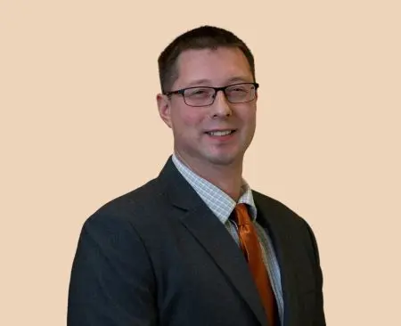 man smiling wearing black suit jacket over a light blue button-up shirt with orange tie