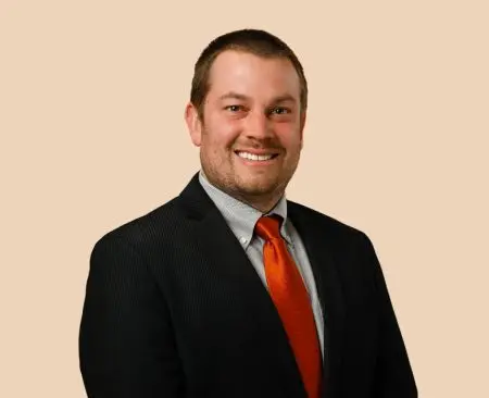 man smiling wearing black suit jacket over a gray button-up shirt with orange tie