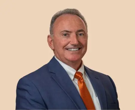 man smiling wearing a blue suit jacket over a white button-up shirt with an orange tie