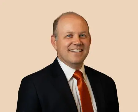 man smiling wearing black suit jacket over a white button-up shirt with orange tie